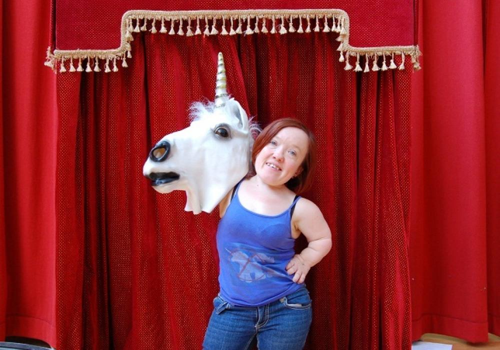 A woman hands against a red curtain backdrop holding one hand on her hip, the other hand is holding up a unicorn head. She is smiling.