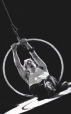 A stark black and white image of a man grapping a large ring that's connected to a wire leading to the sky. Half of his body in laying on the ground, while his torso is encircled by the hanging ring.