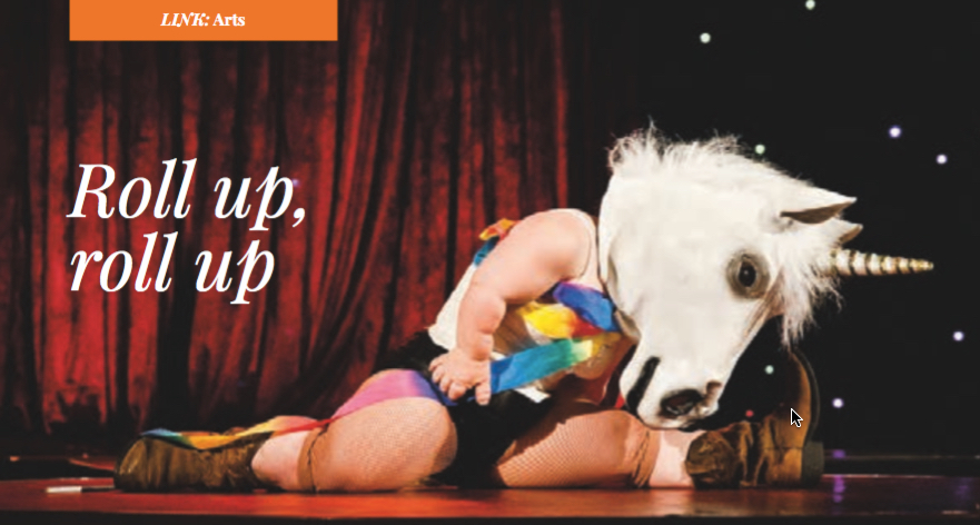 A little woman doing the splits on a stage with a realistic unicorn head covering her face and a colourful ribbon wrapped around her. Bold text on the left hand side reads: Roll up, roll up